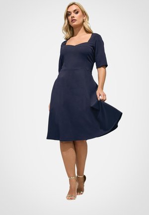 Blonde Frau trägt ein marineblaues knielanges Kleid mit Ellenbogenärmel und quadratischem Ausschnitt, hält beim Stehen die Seite des Kleides fest.