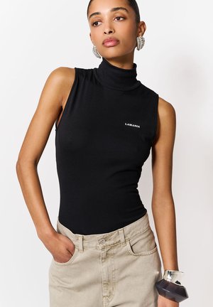 Kobieta ubrana w czarny bezrękawnik z golfem z logo "LAMANIA" oraz beżowe spodnie z wysokim stanem, ozdobiona dużymi kolczykami i wyrazistą bransoletką.