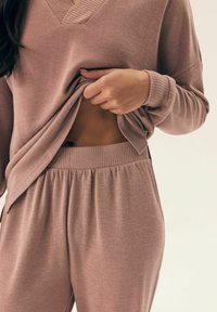 Jasnoróżowy, teksturowany sweter robiony na drutach z dekoltem w kształcie litery V i luźnym krojem, w zestawie z dopasowanymi spodniami na elastycznej talii. Odsłania skórę w okolicy brzucha.