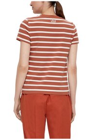 Braun-weiß gestreiftes T-Shirt mit kurzen Ärmeln, runder Ausschnitt und einem kleinen gestickten Design auf dem oberen Rücken. Kombiniert mit orangefarbenen Hosen.