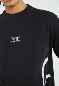 Svart bomullströja med en vit logotyp som har horisontella ränder och en svängd grafik på sidan, rund halsringning och långa ärmar.