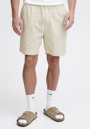 Pantaloni scurți bej cu talie elastică și șnur. Purtați cu ciorapi sport albi și sandale maro cu talpă plată. Textură de țesătură fină.