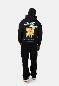 Felpa nera con una grafica colorata di frutta sulla schiena, che presenta pesche arancioni e foglie verdi. Abbinata a pantaloni cargo neri e sneakers.