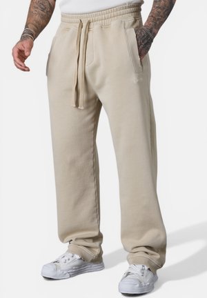 Beige Sweatpants mit Kordelzug, getragen von einer tätowierten Person mit weißen Sneakers, Hände entspannt, eine in der Tasche, stehend auf weißem Hintergrund.