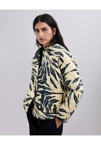 Chaqueta acolchada en crema con rayas de tigre negras, con un escote redondo, botones frontales y un bolsillo de parche. Textura suave y diseño cálido.