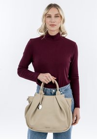 Beige leather handbag with a soft, slouchy design, double top handles, and a metallic tag. Model wears a burgundy long-sleeve sweater and blue jeans.