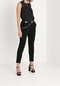 Top senza maniche nero con pois bianchi e scollatura in pizzo, abbinato a pantaloni neri aderenti, cintura a catena argento e sandali neri con tacco alto.