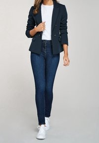 Blazer doppiopetto blu navy, t-shirt bianca, jeans scuri, sneakers bianche. Tessuto liscio, vestibilità sartoriale, gambe dei pantaloni slim, hardware minimale.