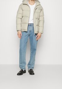 Ljusgrå pufferjacka med huva, med stora kviltade sektioner, dragkedjestängning och sidofickor, bärs över en vit t-shirt och ljusa jeans.