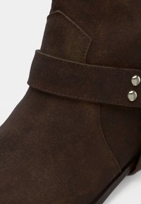 Κοντινό πλάνο σκούρου καφέ δερμάτινου μποτάκι αστραγάλου με διακοσμητικό λουρί και δύο ασημένιες καρφίτσες στο πλάι.