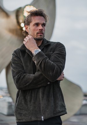 Dunkelgrüne Cordjacke mit Reißverschluss und Kragen, getragen von einer Person. Der Hintergrund zeigt ein großes skulpturales Element.