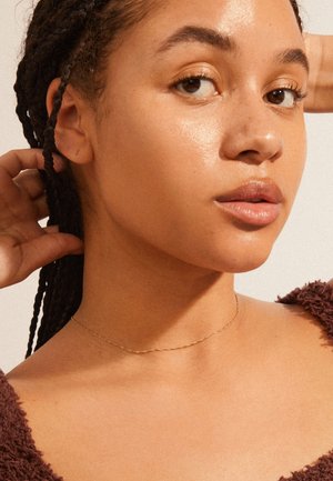 Nahaufnahme einer Person mit Zöpfen, die eine zarte goldene Halskette trägt. Die Haut wirkt glatt und strahlend, mit natürlichem Make-up.