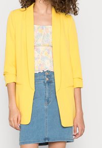 Žena v jasně žlutém blejzru přes pastelový květinový top a modré džíny s vysokým pasem a dvojitými knoflíky.