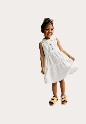 Uśmiechnięta dziewczyna w białej sukience na ramiączkach, żółtych sandałach i czerwonych okularach przeciwsłonecznych na głowie, trzymająca sukienkę jedną ręką.