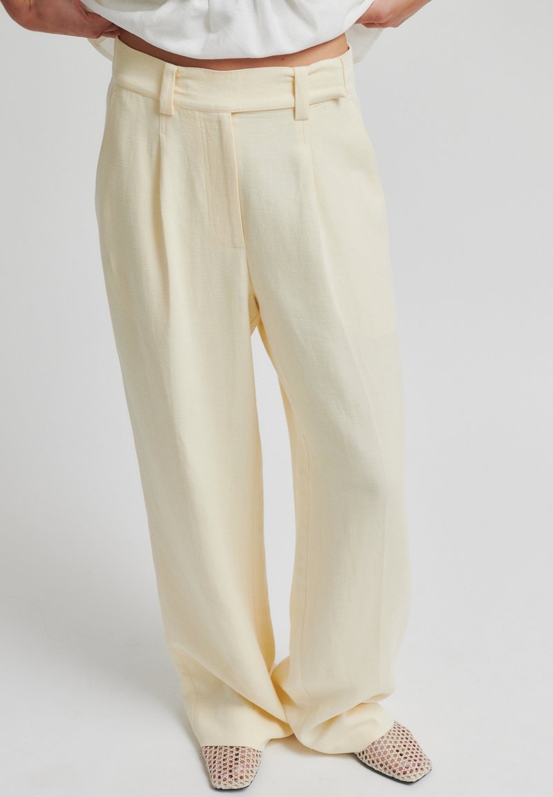 Pantalon large crème avec passants de ceinture porté avec des chaussures blanches en mesh à bouts carrés sur un fond clair uni.