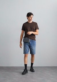 T-shirt marrone a girocollo, pantaloni shorts in denim azzurro chiaro, scarpe nere e calze nere con una borsa in vita grigia. In piedi su un pavimento grigio contro un muro semplice.