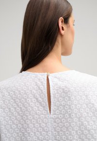 Witte blouse met een textuur van bloemenpatroon, voorzien van een opening met sleutelgat aan de achterkant, vastgezet met een decoratieve knoop. Gemakkelijke stof, casual ontwerp.