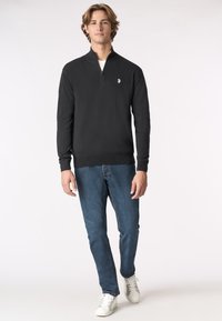 Maglione nero con zip e collo alto, maniche lunghe, texture a maglia, dettaglio del logo discreto sul petto, abbinato a jeans blu e sneakers bianche.