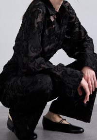 Tenue en superposition noire transparente avec texturation florale ; manches larges et pantalons amples. Chaussures noires à petit talon, les mains reposant sur les genoux.