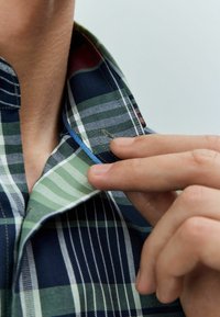 Herrenhemd mit Karomuster in Marineblau, Grün und Weiß. Verfügt über einen kontrastierenden Innenkragen mit blauen Akzenten. Hand, die eine Verschlussdetails anpasst.