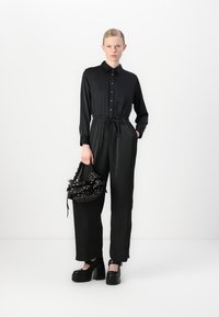 Zwarte jumpsuit met een kraagomslag, knopen aan de voorkant en een trekkoord in de taille. Heeft lange mouwen en een brede broekspijp. Zwarte platform schoenen en tas.