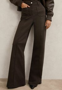 Hnedé džíny s vysokým pásom a zvonovými nohavicami, s prednými vreckami, hladkou textúrou, čistými líniami a jemným prešívaním. Kombinované s priliehavou tmavou bundou.