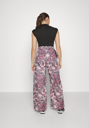 Vrouw staand met de rug naar de camera, draagt een mouwloos zwart topje en broek met een wijd uitlopende pijp in roze, zwart en wit patroon, haar in een paardenstaart.