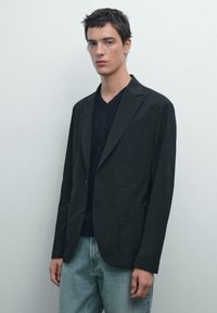Schwarzer Blazer mit Reverskragen, zwei Taschen vorne und einem einfachen Knopfverschluss, getragen über einem dunklen V-Ausschnitt-Shirt und hellen Jeans.