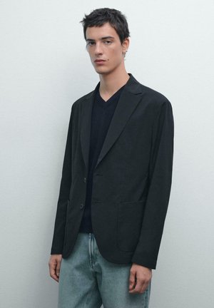 Schwarzer Blazer mit Reverskragen, zwei Taschen vorne und einem einfachen Knopfverschluss, getragen über einem dunklen V-Ausschnitt-Shirt und hellen Jeans.