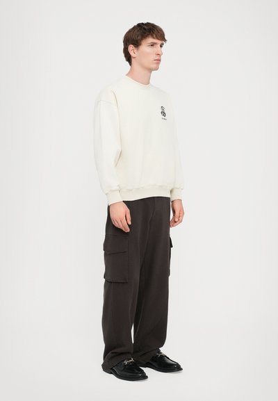 Cremefarvet sweatshirt med ribstrikkede manchetter og kant, prydet med et lille sort logo. Sammen med løstsiddende mørke cargo-bukser med sidelommer.