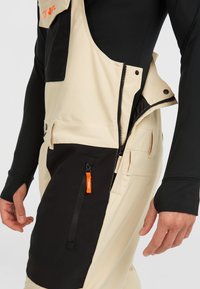 Pantalones de peto impermeables en beige y negro con un bolsillo con cremallera, cierre de botón lateral y acento naranja en el tirador de la cremallera. Textura suave.