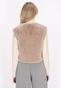 Faux-Fur-Weste in hellbeige mit weicher Textur, hochgeschlossen und ärmellos, getragen über einem cremefarbenen Langarmshirt und grauen Hosen.