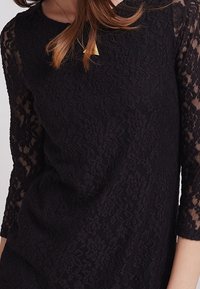 Svart spetstopp med trekvartsärmar, intrikata blommönster och rund halsringning med ett mjukt tyg underlager.