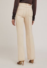 Pantalons évasés beiges avec une taille haute, des coutures marquées, deux poches arrière et un logo visible sur la bande de taille, associés à des chaussures à talons hauts.