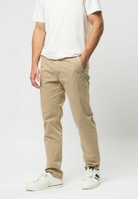 Vyras dėvi smėlio spalvos kelnes, baltus sportinius batelius su mėlynomis ir raudonomis juostelėmis bei baltą trumpomis rankovėmis marškinį, stovintis su viena ranka kišenėje.