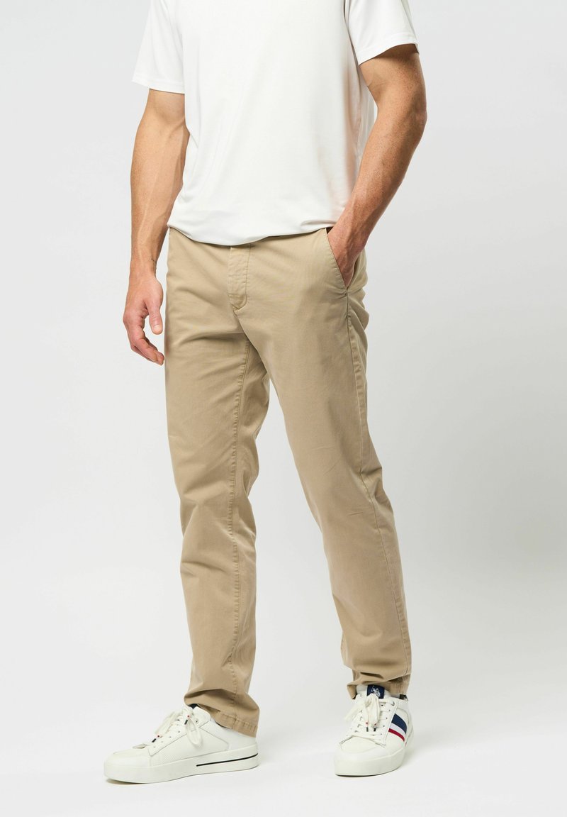 Vyras dėvi smėlio spalvos kelnes, baltus sportinius batelius su mėlynomis ir raudonomis juostelėmis bei baltą trumpomis rankovėmis marškinį, stovintis su viena ranka kišenėje.