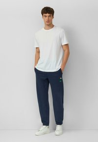 Młody mężczyzna w białej koszulce z okrągłym dekoltem, granatowych spodniach dresowych z zielonym nadrukiem żółwia oraz białych sportowych butach, stojący na tle jasnej, gładkiej ściany.