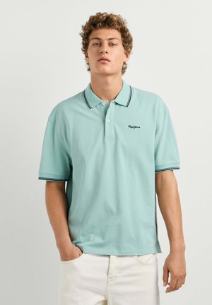Junger Mann mit lockigem Haar, der ein hellblaues Poloshirt mit schwarzem Rand und weiße Hose trägt, steht mit einer Hand in der Hosentasche.