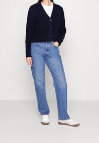 Marinblå kortärmad cardigan med ribbat mönster, tre knappar och två framfickor, ihop med ljusblå raka jeans och vita sneakers.