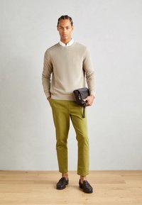 Pull en tricot beige, pantalons ajustés verts, sac bandoulière noir et mocassins foncés. Texture lisse, design ajusté et détails minimaux.