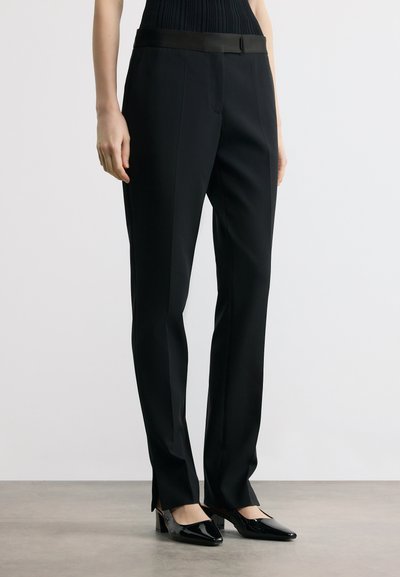 Femme portant un pantalon noir sur mesure avec un détail ceinture en satin, assorti à des chaussures noires en cuir verni à petit talon, debout sur un sol gris.