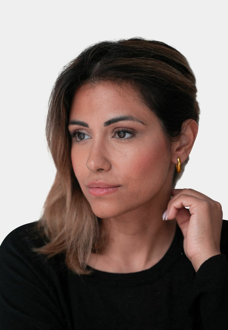 Boucles d'oreilles en or, cheveux longs avec des mèches plus claires, portant un haut noir. Peau lisse, sourcils visibles et expression faciale naturelle.