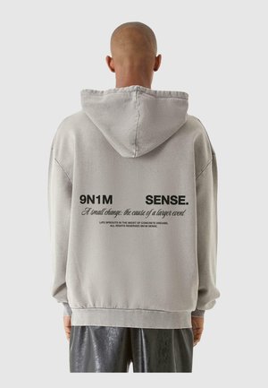 Sudadera gris de gran tamaño con capucha, que presenta texto negro en la parte posterior. El material parece suave con un diseño estructurado y puños acanalados.