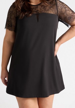 Czarna sukienka z przezroczystymi koronkowymi krótkimi rękawami i dekoltem, noszona przez stojącą osobę z jedną ręką na biodrze na gładkim tle.