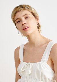 Robe blanche sans manches avec des bretelles larges, décolleté carré et taille à cordon. Texture lisse avec une coupe décontractée, design minimal.