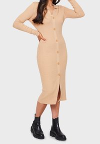 Robe midi en maille côtelée beige avec manches longues, fermeture à boutons sur le devant et fente sur le côté. Association avec des bottines noires.