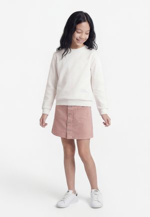 Dziewczyna z długimi ciemnymi włosami ma na sobie białą bluzę, różową spódnicę na guziki i białe trampki, uśmiecha się i spogląda w dół.