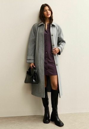 Un manteau en laine gris avec de grands boutons, superposé sur une robe ajustée prune. Des bottes noires jusqu'aux genoux et un sac à main noir complètent le look.