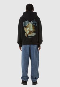 Svart hettegenser med grafisk design av duer og tekst på baksiden, med en avslappet passform. Kombinert med løstsittende blå jeans.