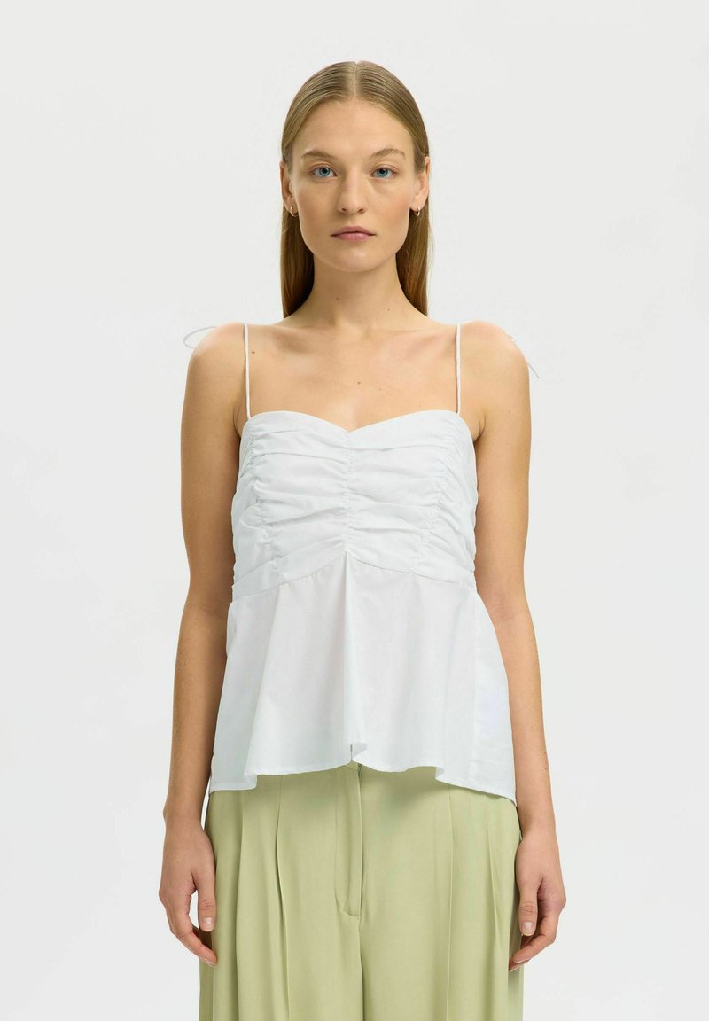 Jeune femme aux longs cheveux raides porte un haut blanc froncé sans manches et un pantalon plissé vert clair, debout face à l'avant.